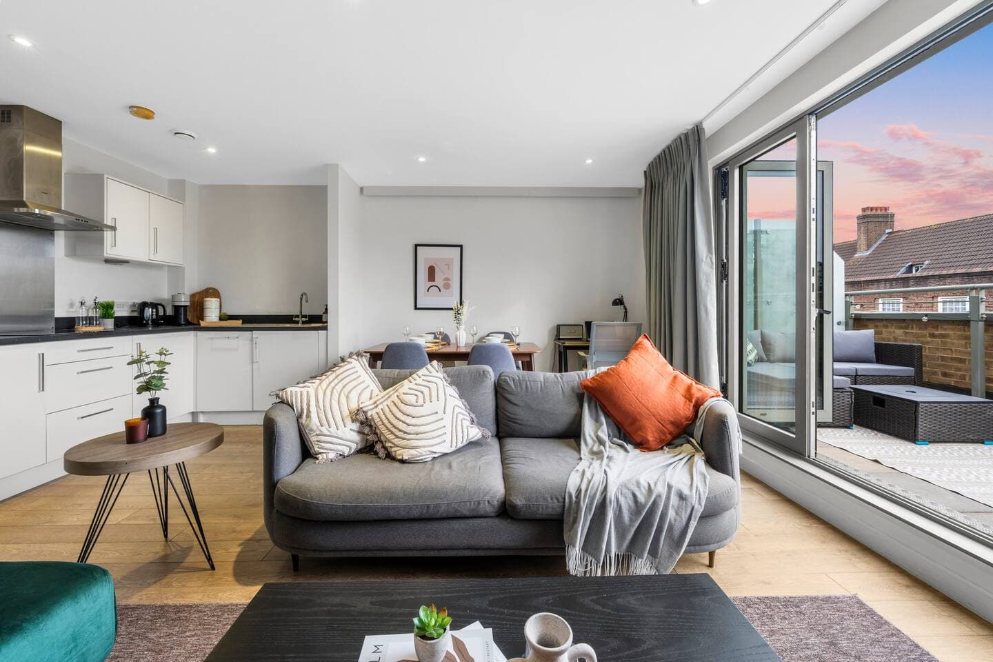 Modern open-plan living space with a fully equipped kitchen, dining area, and comfy sofa. Large glass doors lead to a private balcony with outdoor seating and sunset views, creating a bright, airy setting perfect for relaxing or entertaining.