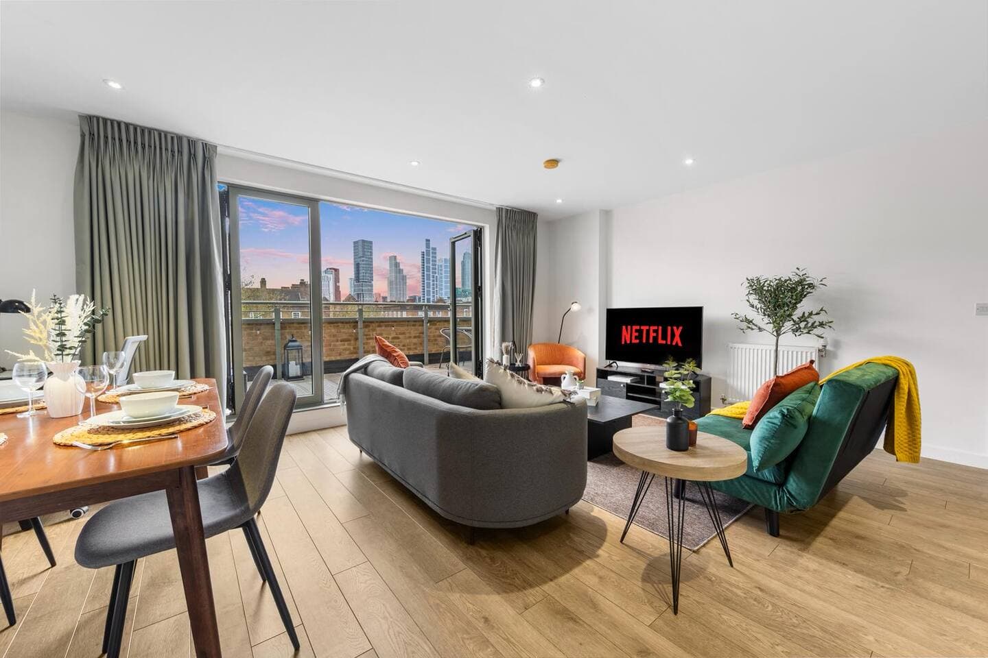 modern open-plan living and dining space with doors opening to a private balcony and skyline views. Features warm wood floors, a cozy gray sofa, green accent chair, smart TV, and a dining table set for four, filled with natural light.